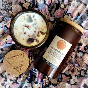 May include: A brown glass candle jar with a cork lid and a label that reads "Full Moon Ritual Clove Coconut Amber Vanilla" with a description of the candle's benefits. The candle is surrounded by a floral fabric.