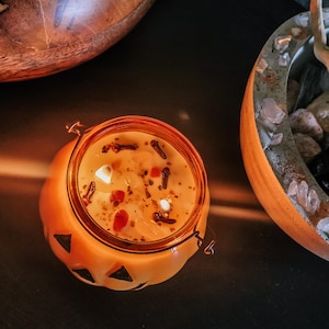 May include: A lit, orange-colored candle in a pumpkin-shaped jar with a metal clasp. The candle wax is speckled with spices and small crystals. The jar has black triangle cutouts. A wooden bowl is in the background.
