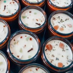 May include: Close-up of multiple amber glass jars filled with white wax and various natural ingredients, including small crystals, herbs, and spices. The jars are arranged in a circular pattern, creating a visually appealing and aromatic display.