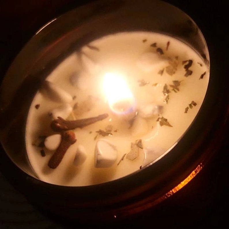 May include: A white candle with a flame burning in the center. The candle is in a clear glass jar with a brown rim. The candle has small pieces of herbs and crystals embedded in it.