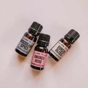May include: Three small brown glass bottles with black caps and white labels. The labels have black text that reads "Woodland Spice", "Birch Bliss", and "Smoked Rose".