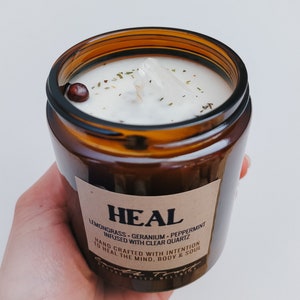 May include: A brown glass candle jar with a white label that reads "HEAL" and "LEMONGRASS - GERANIUM - PEPPERMINT INFUSED WITH CLEAR QUARTZ". The label also says "HAND CRAFTED WITH INTENTION TO HEAL THE MIND, BODY & SOUL".
