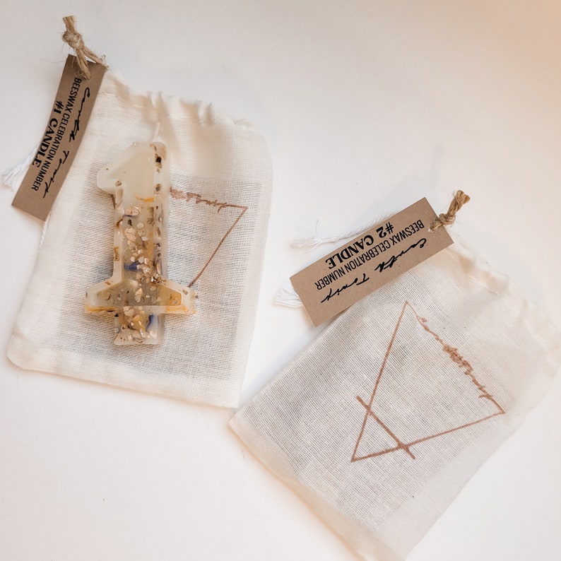 May include: Two small white fabric bags with brown paper tags. The bags contain beeswax candles. The tags have the text "#1 CANDLE BEESWAX CELEBRATION NUMBER" and "#2 CANDLE BEESWAX CELEBRATION NUMBER".