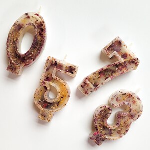 May include: Four white birthday candles with embedded dried flowers and gold glitter. The candles are shaped like the numbers 0, 5, 7, and 3.