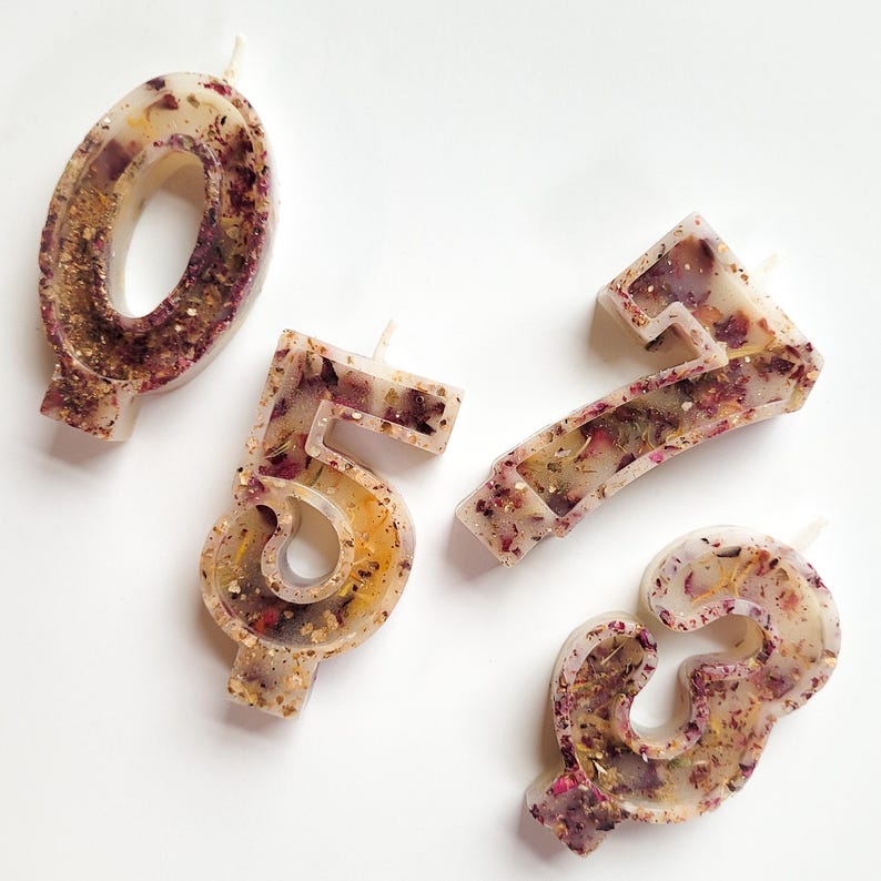 May include: Four white birthday candles with embedded dried flowers and gold glitter. The candles are shaped like the numbers 0, 5, 7, and 3.