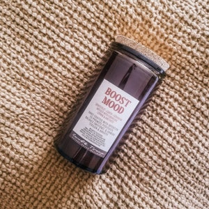May include: A dark brown glass candle jar with a cork lid. The label reads "Boost Mood" and lists grapefruit, lemon, geranium, carnelian, citrine, and amethyst. The candle is hand-crafted with intention to promote positivity, balance emotions, and unwind tension and anxiety.