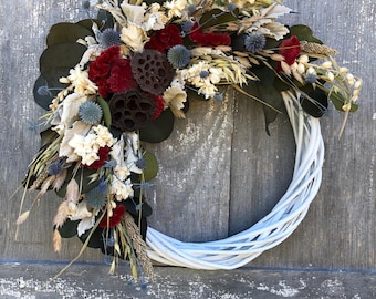 May include: A white woven wreath adorned with dried flowers and foliage. The arrangement includes burgundy, cream, and blue accents, with dark green eucalyptus leaves. The wreath is displayed against a weathered wooden background, creating a rustic aesthetic.