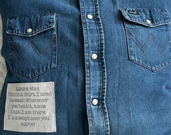 Może przedstawiać: Poduszka z niebieskiego dżinsu z kieszenią i guzikami. Poduszka ma wszyty biały materiałowy łatkę z tekstem "Laura Mae, This is a shirt I used to wear, Whenever you hold it, know that I am there. I'l always love you. Aaron".
