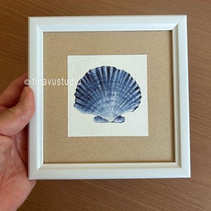 May include: A square white frame containing a watercolor painting of a single blue scallop shell on a cream-colored background. The artwork is centered within a tan mat board. The text tinavustudio is printed in white on the left side.