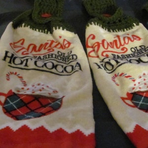 May include: Two white kitchen towels with green crocheted tops. The towels feature red and green plaid mugs with whipped cream and candy canes. The text on the towels reads "Santa's Old Fashioned Hot Cocoa" and "Grinch's Old Fashioned Hot Cocoa".