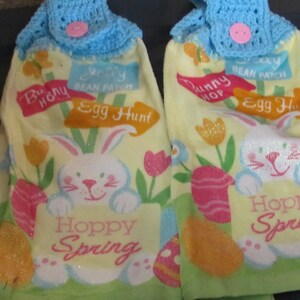 May include: Two yellow kitchen towels with a bunny rabbit design and the text "Bunny Hop Bean Patch Egg Hunt" and "Hoppy Spring". The towels have blue crocheted tops with a pink button.