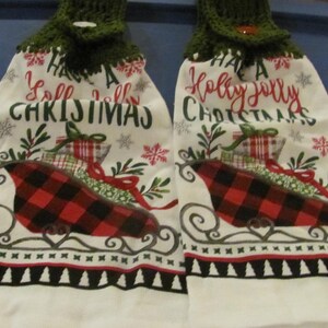 May include: Two white kitchen towels with a red and black plaid design and a green crocheted top. The towels have the text "Have a Holly Jolly Christmas" and "Have a Y'all Y'all Christmas" printed on them.