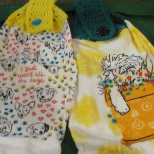 May include: Two white kitchen towels with crocheted tops. One towel has a yellow crocheted top with a blue button and features a dog print with the text "I just want all the dogs". The other towel has a green crocheted top with a brown button and features a white dog in a yellow pot.
