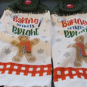 May include: Two white kitchen towels with red and white checkered borders. Each towel features a gingerbread man with a red and white striped scarf, snowflakes, and the text "Baking Spirits Bright". The top of each towel has a green crocheted border with a red button.