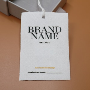 Puede incluir: Una etiqueta rectangular blanca con las palabras "BRAND NAME" en letras negras grandes, con "OR LOGO" debajo. La etiqueta incluye líneas para "Any text/color/design" y "Handwritten Notes:" en la parte inferior. Una cuerda blanca está adjunta.