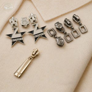 May include: A collection of metal zipper pulls and zipper heads in silver and gold tones. Some pulls feature a star design with the text "ZINAPUNK". The items are displayed on a cream-colored fabric.