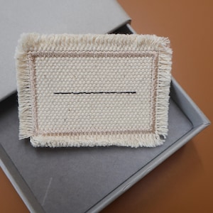 May include: A rectangular, off-white woven coaster with a black horizontal line and fringed edges. The coaster has a gold border and is presented inside a gray box, ideal for home decor or as a gift.