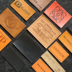 Puede incluir: Surtido de parches de cuero con varios nombres de marca y logotipos, incluyendo Playboy, Versace, Levi Strauss & Co., Burberry y Calvin Klein Jeans. Los parches son de color marrón, negro y naranja.