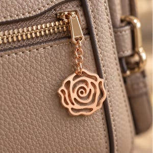 May include: A taupe-colored bag with a textured, pebbled surface featuring a gold-toned metal zipper. A decorative rose-shaped charm hangs from the zipper pull by a small chain, matching the metallic finish of the zipper hardware and the bag's trim.
