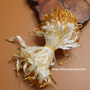 Pode incluir: Um conjunto de fitas bege com alfinetes dourados em forma de cabaça. As fitas são amarradas com uma faixa amarela. Os alfinetes têm um acabamento dourado. O texto "Beige ribbon gold gourd pin" é visível.