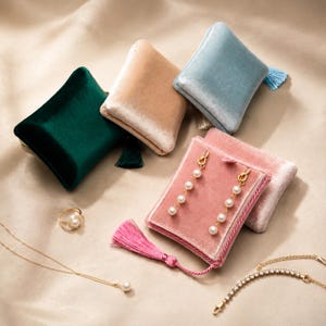 Könnte beinhalten: Eine Sammlung quadratischer Schmuckbeutel aus Samt in Dunkelgrün, Beige, Hellblau und Rosa, jeweils mit einer passenden Quaste. Ein Paar goldene Ohrhänger mit weißen Perlen, ein goldener Ring mit Perle, eine Goldkette und ein Armband sind zu sehen.