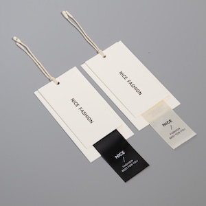May include: Two rectangular white clothing tags with the text "NICE FASHION" printed in black. One tag has a black label with "NICE / FASHION BEST FOR YOU". The other tag has a transparent label with the same text. Both tags have a string attached.
