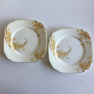 May include: Two square, cream-coloured plates with gold rims. Each plate has a decorative design of yellow flowers and a gold, leaf-like pattern. The plates are likely part of a vintage or antique dinner service.