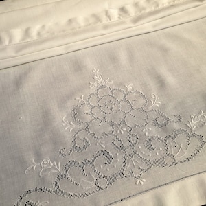 Puede incluir: Funda de almohada de lino blanco con un delicado diseño floral bordado. El bordado presenta una flor grande con flores y hojas más pequeñas que la rodean.