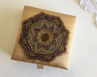 May include: A gold satin box with a beaded design in shades of brown, gold, and purple. The box has a small loop on the front.