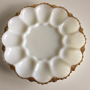 May include: A round white milk glass egg plate featuring twelve individual oval indentations arranged in a circular pattern. The scalloped outer edge of the serving dish is finished with a metallic gold trim that highlights the curved rim.