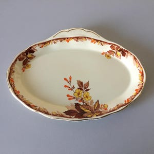 May include: An oval ceramic serving platter with a cream-colored base and a scalloped edge. The rim features a decorative border of brown and orange autumn leaves. A central floral motif displays yellow blossoms and brown foliage on the surface.