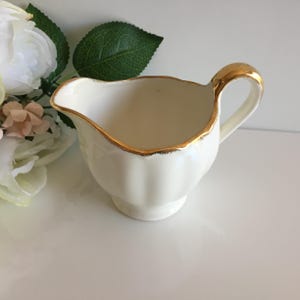 May include: A cream-coloured ceramic creamer with a gold rim and handle. The creamer has a classic design with a slightly fluted body and a small, elegant spout. The gold trim adds a touch of sophistication to this vintage-style serving piece.