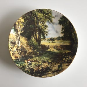 May include: A decorative plate featuring a pastoral landscape scene with trees, a stream, and sheep. The plate has a gold rim and depicts a detailed painting of a countryside setting with figures and a dog. The colors include greens, browns, and blues.