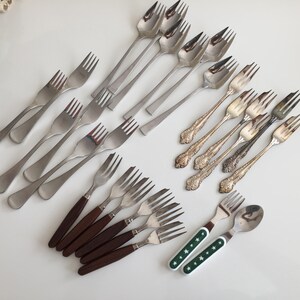 Puede incluir: Un surtido de tenedores y cucharas vintage en varios estilos. La colección incluye tenedores plateados, tenedores con mangos de madera y un juego de tenedor y cuchara con mangos verdes y blancos con estampado de estrellas.