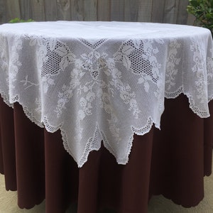 May include: White lace tablecloth with a floral pattern, draped over a brown round table.