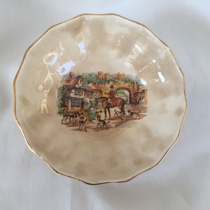 May include: A decorative, scalloped-edge bowl with a cream-colored background and a gold rim. The center features a colorful illustration of a hunting scene with people, dogs, and a horse. A charming piece for display.