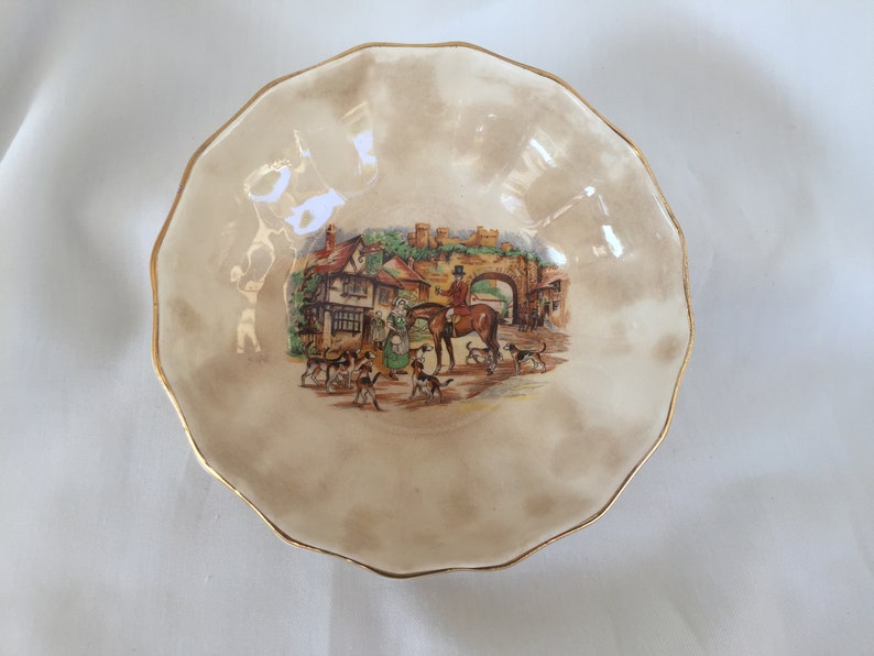 May include: A decorative, scalloped-edge bowl with a cream-colored background and a gold rim. The center features a colorful illustration of a hunting scene with people, dogs, and a horse. A charming piece for display.