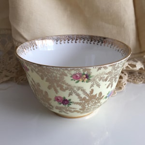 May include: A vintage porcelain bowl. The pale yellow exterior is adorned with gold filigree and small floral bouquets in pink, purple, and blue. The interior is white, and the rim features a gold pattern. A classic piece for serving.