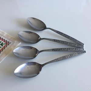 Puede incluir: Cuatro cucharas de acero inoxidable con cuencos ovalados y patrones florales decorativos en los mangos. Las cucharas están dispuestas sobre una superficie blanca, con un pequeño paño bordado en la esquina.