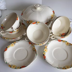 Könnte beinhalten: Cremefarbenes Vintage-Porzellanservice mit floralen Akzenten. Das Set umfasst eine Servierschüssel mit Deckel, Schalen, Tassen und Untertassen. Die Ränder der Gerichte sind mit gelben und orangen Blumen verziert.