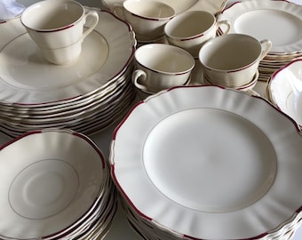 May include: A collection of cream-colored vintage dishware with a scalloped edge and a burgundy trim. The set includes multiple plates, saucers, and teacups, arranged in stacks and groupings. The dishes have a delicate, classic design.