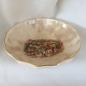 May include: A small, cream-colored decorative dish with a scalloped edge and gold trim. The center features a detailed illustration of a hunting scene with people, dogs, and a horse. A charming vintage piece.