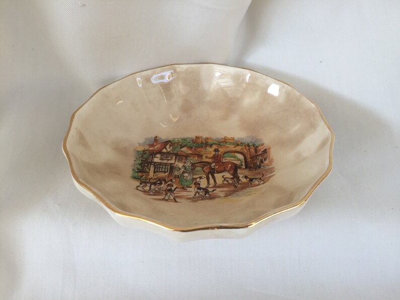 May include: A small, cream-colored decorative dish with a scalloped edge and gold trim. The center features a detailed illustration of a hunting scene with people, dogs, and a horse. A charming vintage piece.