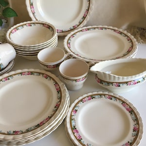 May include: A vintage cream-coloured china set featuring plates, bowls, cups, and a gravy boat. Each piece has a scalloped edge and a border of pink and blue roses with gold trim. The china set is decorated with a floral design.