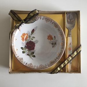 May include: A white porcelain bowl with a floral rose design in red and orange, and gold trim, presented in a gold box with a matching ice cream scoop. The set is tied with a black and gold ribbon, ideal for gifting.