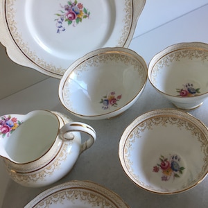 Puede incluir: Un juego de té vintage con un diseño floral. El juego incluye una tetera, una jarra de crema, un azucarero, tazas de té y platillos. Las piezas son blancas con ribetes dorados y presentan un delicado patrón floral.