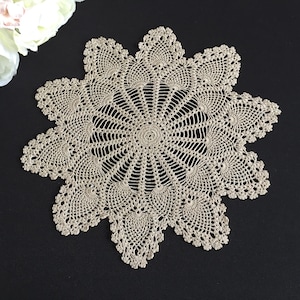 Könnte beinhalten: Handgefertigte, beige Häkelspitze mit floralem Design. Die Spitze hat ein zentrales kreisförmiges Muster mit radialen Linien und einem gewellten Rand. Die Spitze liegt auf einer schwarzen Oberfläche, mit weißen Blumen in der oberen linken Ecke.