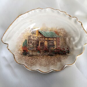 May include: A white ceramic trinket dish shaped like a seashell with a gold rim. The dish features a painted image of a cottage with a green roof and red flowers.