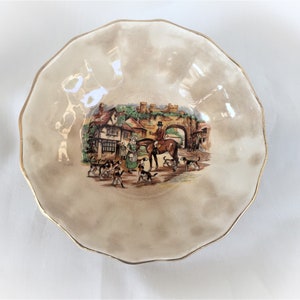 May include: A decorative, scalloped-edge bowl with a gold rim. The bowl features a central illustration of a hunting scene with people, dogs, and a horse. The background is a muted beige color.