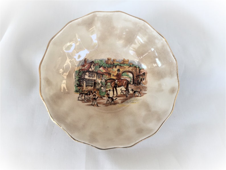 May include: A decorative, scalloped-edge bowl with a gold rim. The bowl features a central illustration of a hunting scene with people, dogs, and a horse. The background is a muted beige color.